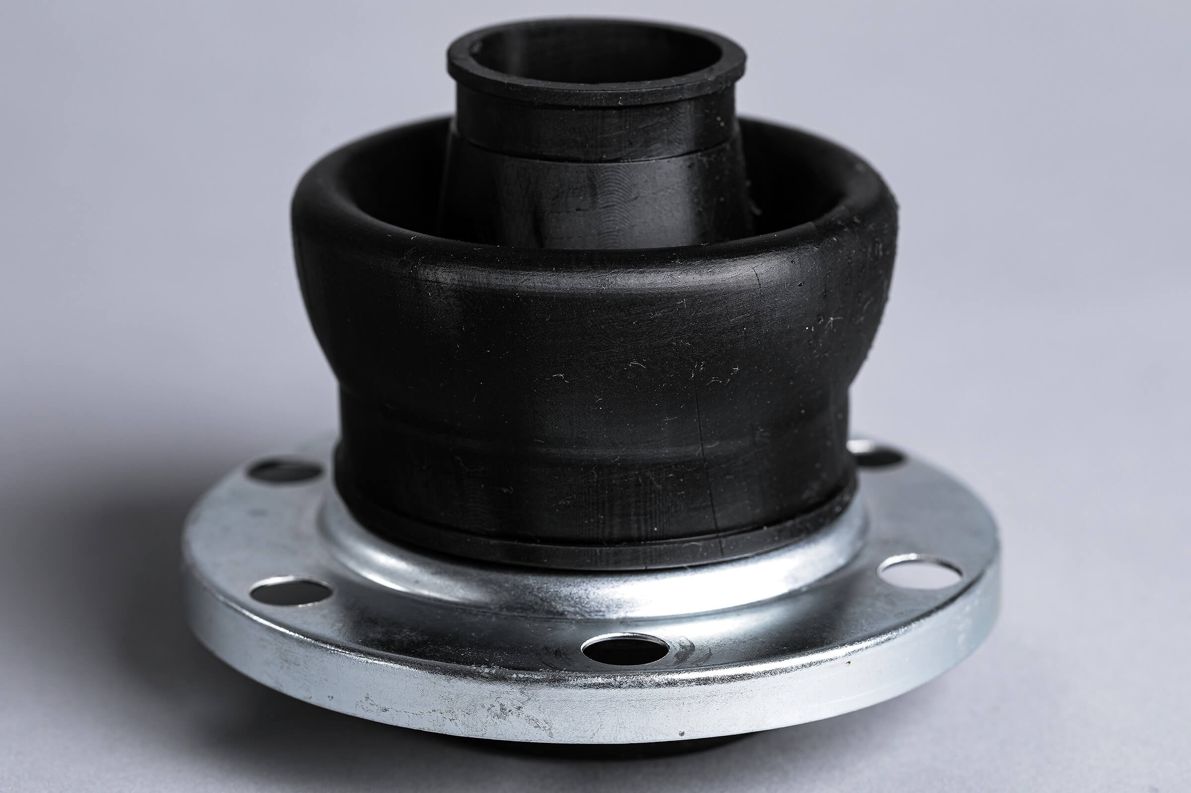 Close-up of a black rubber and silver metal car suspension mount with circular holes, placed against a plain gray background.