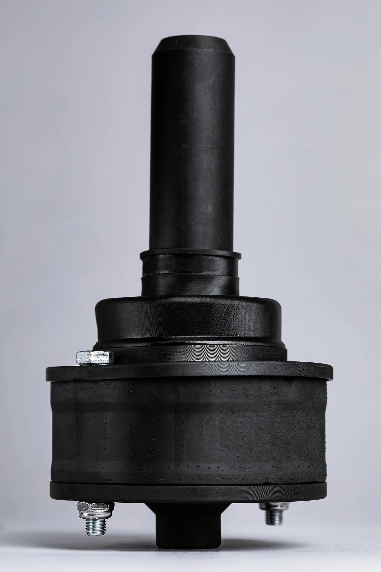 A black mechanical component with cylindrical and round elements, featuring metallic bolts, set against a neutral gray background.
