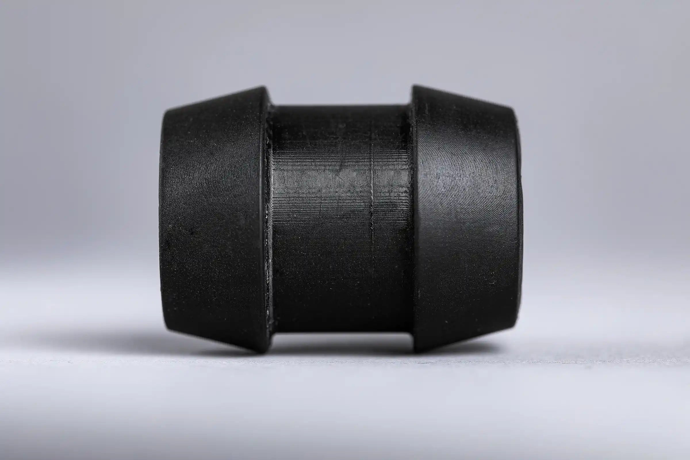 A black, cylindrical rubber grommet with angled ends sits on a smooth, gray surface, casting a soft shadow. The lighting is neutral and even.