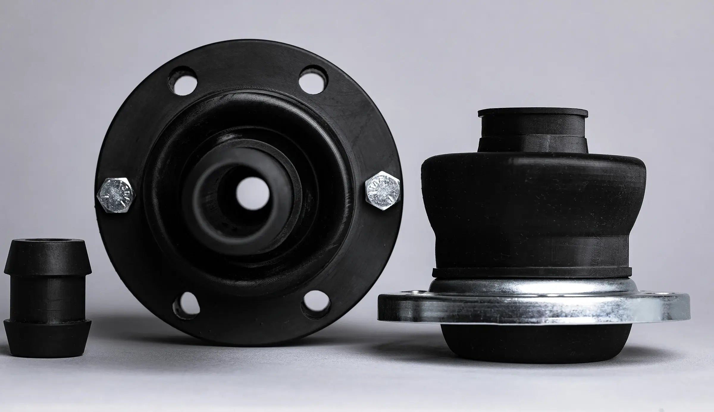 Close-up of two black rubber automotive parts on a gray background, featuring bolts and metal accents. The mood is industrial and precise.