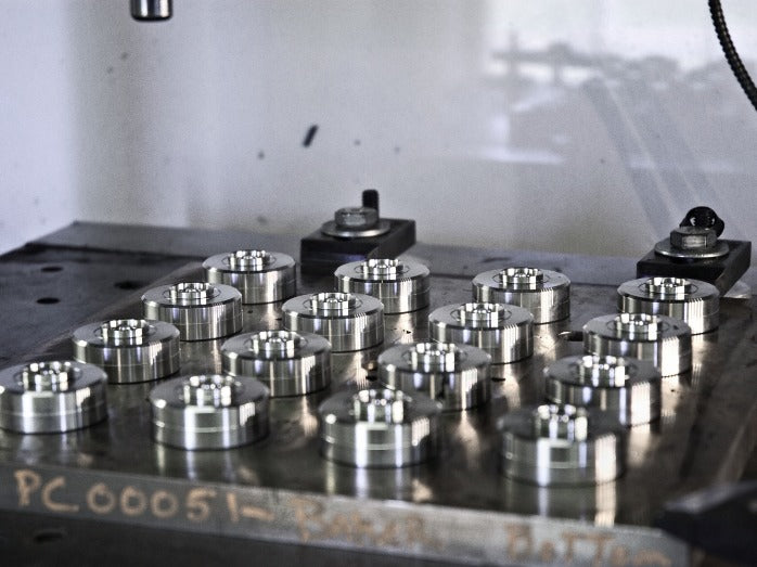 Sixteen shiny metal pieces are arranged in rows on a workshop table. The industrial setting is highlighted by soft lighting and a neutral tone.