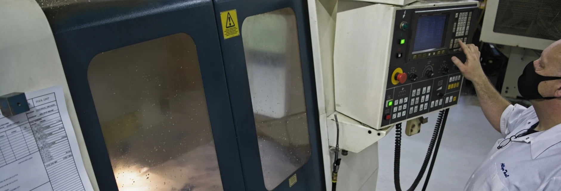 A person operates a CNC machine, pressing buttons on its control panel. The machine's window shows interior machinery. The scene conveys focus and precision.
