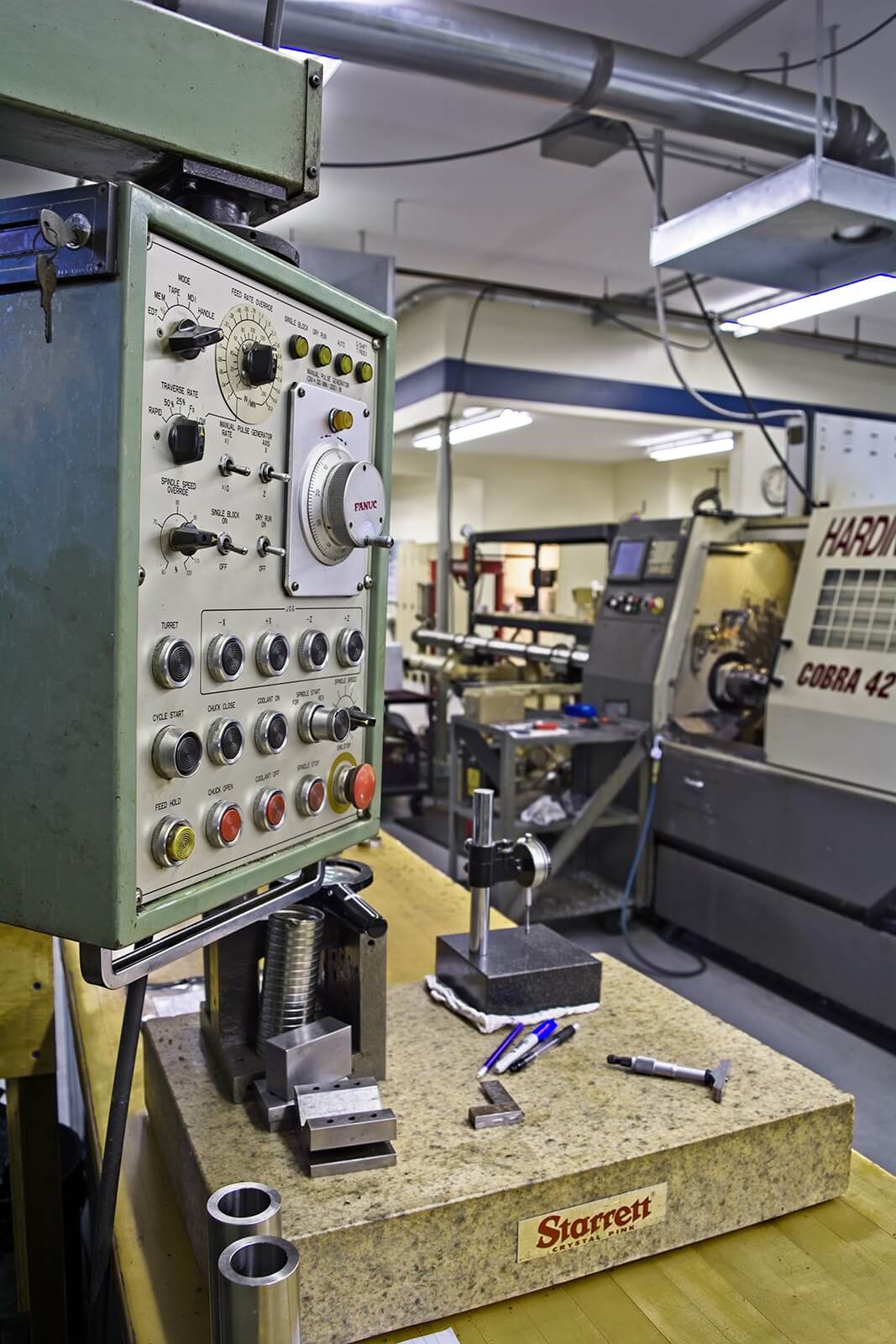 Industrial workshop with a CNC machine and control panel in the foreground. Tools and precision instruments are on a granite surface, suggesting detailed engineering work.