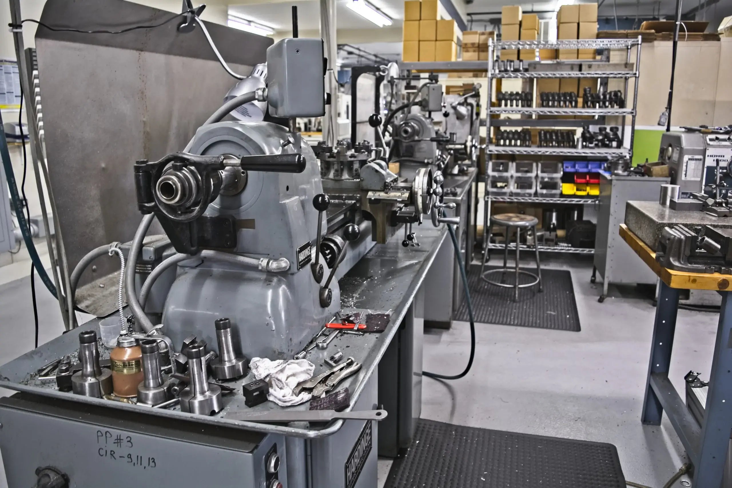 A workshop with industrial machinery, including metal lathes and tools. Shelves hold organized boxes and supplies, creating a busy, functional atmosphere.