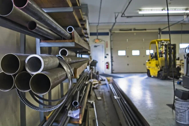 Workshop interior with metal pipes stacked on shelves. A yellow forklift is in the background near a large, closed garage door. Industrial setting.