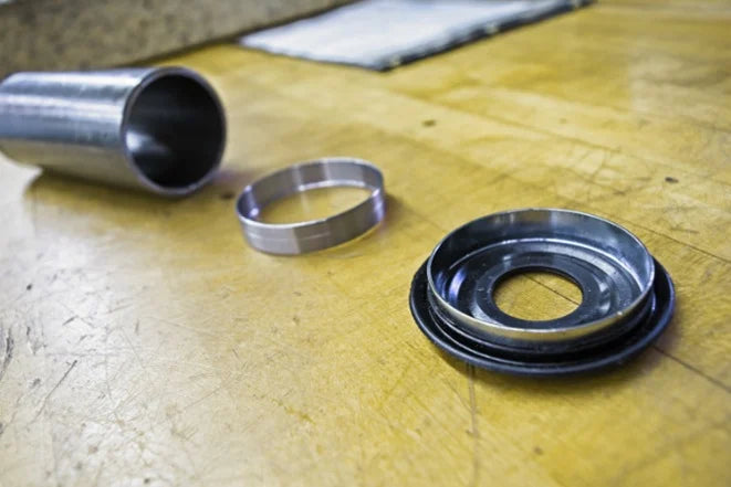 Metal parts are spread on a wooden workbench, including a cylindrical tube, a ring, and a circular assembly. The setting feels industrial and precise.