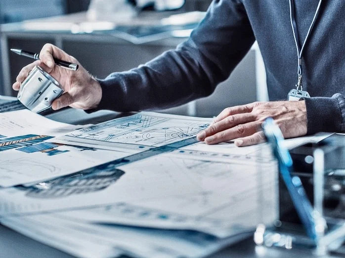 A person reviews architectural plans on a desk, holding a pen. The scene conveys a focused, professional atmosphere, emphasizing precision and design.