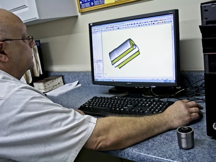 A person in a white shirt is working at a computer with a 3D design of a mechanical part on the screen. The setting is an office with a focused atmosphere.