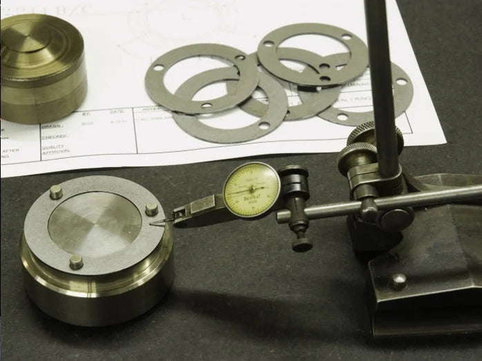 A dial indicator measures a metal disc's precision, with circular gaskets and a technical drawing in the background, suggesting detailed engineering work.
