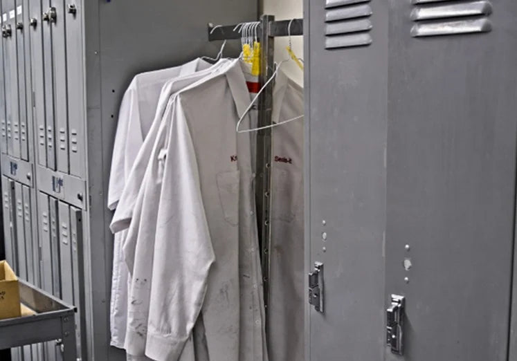 White lab coats hang inside a gray metal locker, flanked by more lockers. The scene feels industrial and utilitarian, conveying a sense of work readiness.