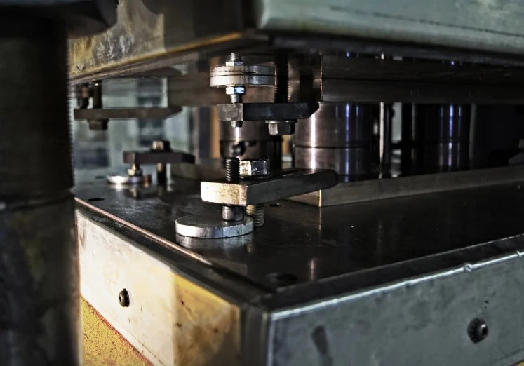 Close-up of an industrial metal press with intricate gears and levers; the scene conveys precision and engineering complexity in a factory setting.