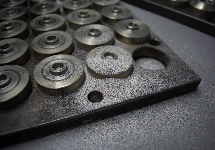 A perforated metal plate holds circular metal components dusted with gray particles, conveying an industrial and orderly atmosphere.