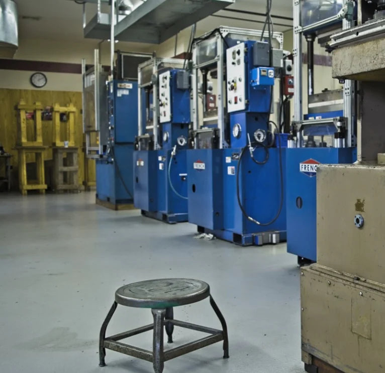 Industrial room with a line of blue hydraulic presses in the background. A single metal stool sits in the foreground. The space is orderly and functional.