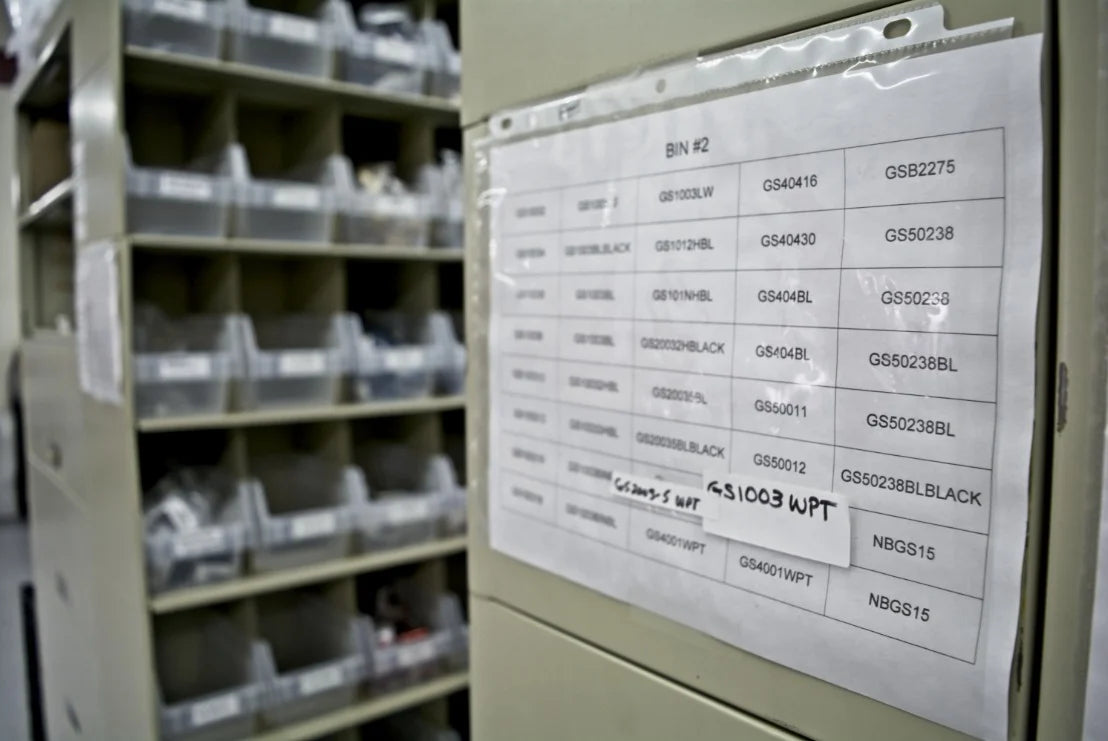 Close-up of a storage area with a labeled chart, "BIN #2," listing codes and items. Shelves with plastic bins are visible in the background. Industrial and organized setting.