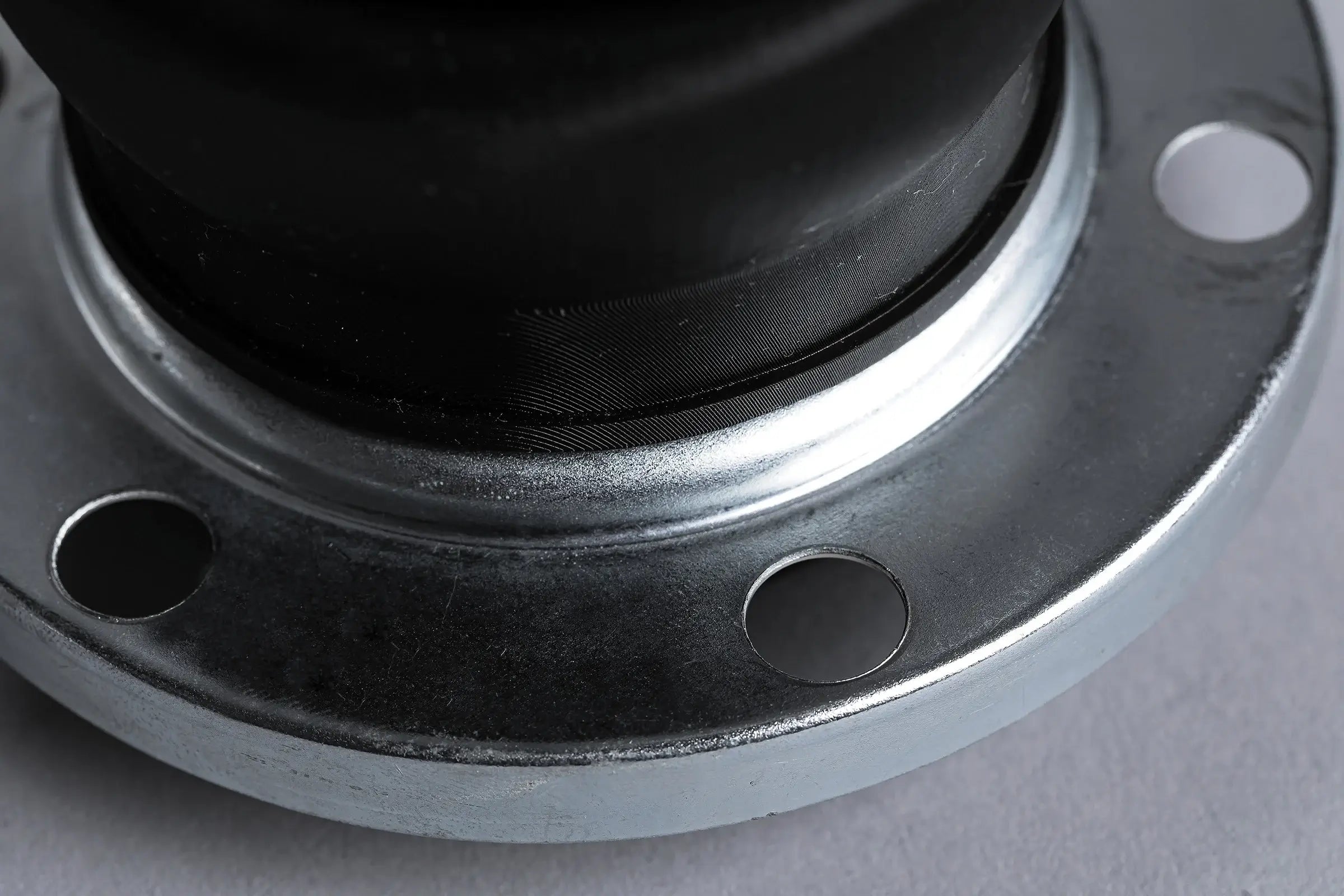 Close-up of a metallic flange with four evenly spaced holes, attached to a black cylindrical component, set against a neutral gray surface.