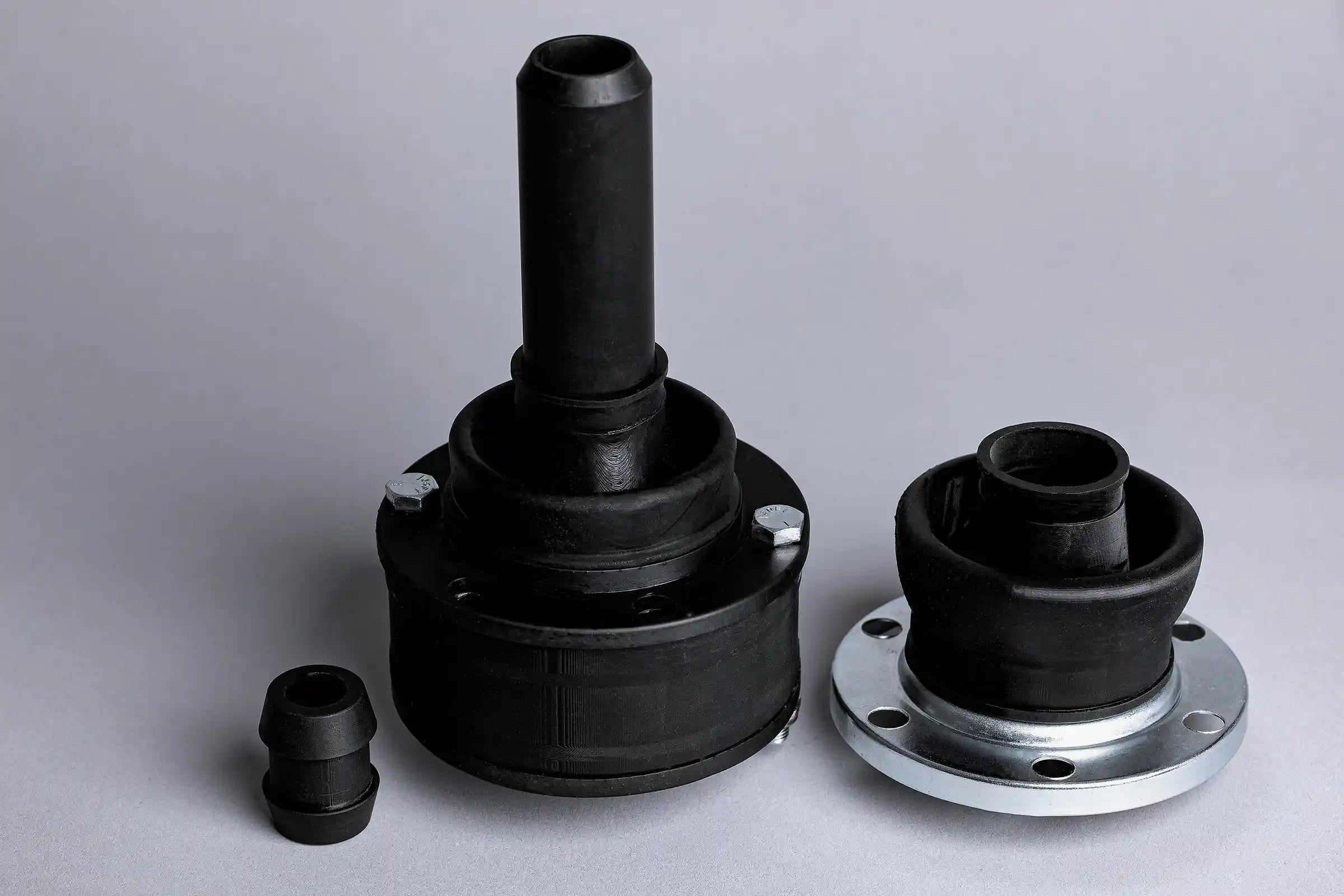 Three black rubber automotive grommets of varying sizes are displayed on a light gray surface. The largest has a metal base with bolt holes.
