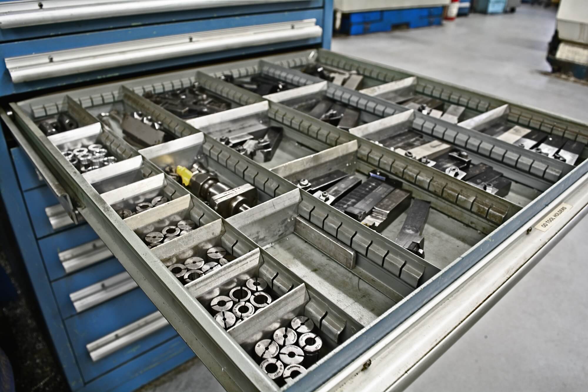 Open blue metal drawer with organized metal tools and parts in compartments. The industrial setting conveys order and efficiency.