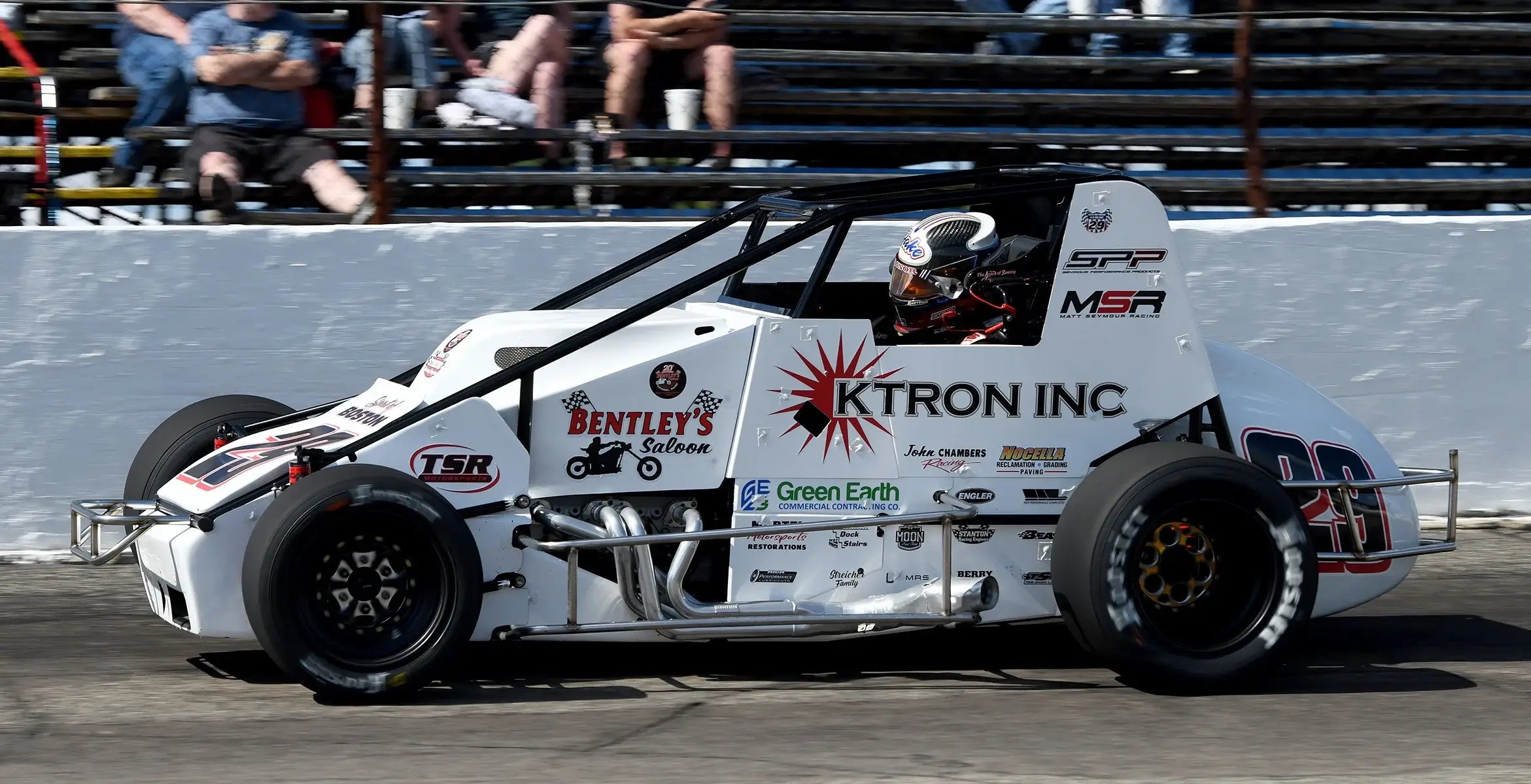 A white sprint car speeds along a racetrack. The car features logos, including "Ktron Inc" and "Bentley's Saloon," with a focused driver inside.