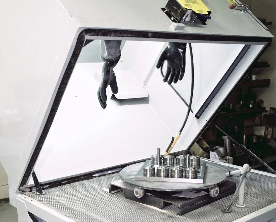 A laboratory glove box with black gloves attached inside. Metallic components are placed on a rotating table, suggesting precision work and safety.